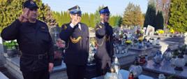 Widok z przodu. Trzech strażaków PSP salutuje na cmentarzu przed grobem. Na drugim planie inne mogiły. 