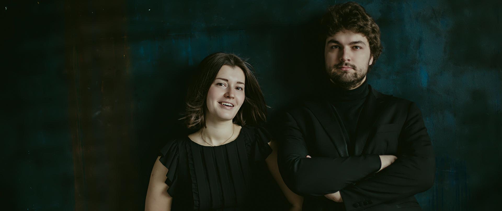 Na zdjęciu kobieta w ciemnych włosach ubrana na czarno. Obok nie mężczyzna w czarnych włosach i brodzie. Mężczyzna ubrany w czarny golf i czarną marynarkę. Stoją na tle ciemnej ściany.