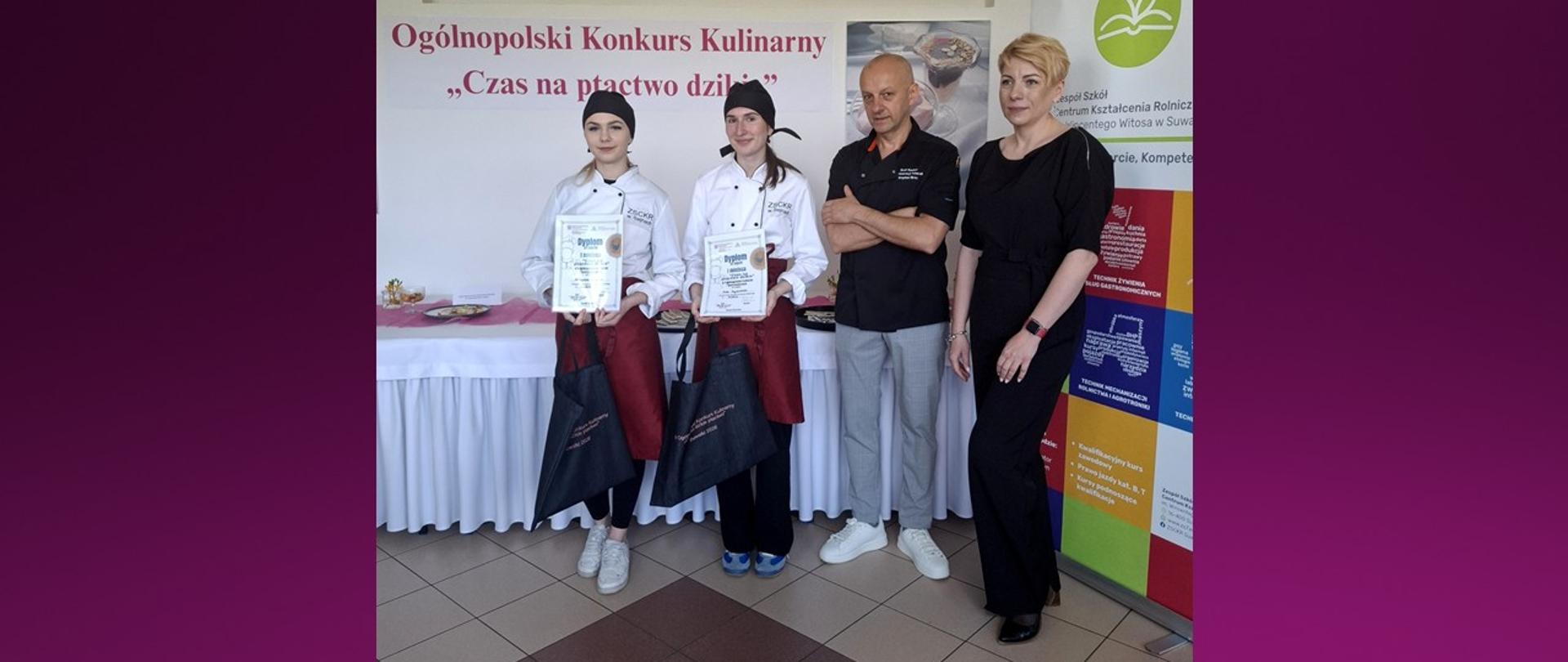 Dwie uczennice ubrane w stroje kucharskie, zwyciężczynie konkursu kulinarnego, pozują do zdjęcia wraz kobietą i mężczyzną.