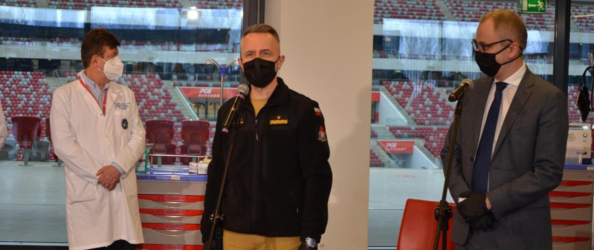 Zdjęcie przedstawia trzech mężczyzn w maseczkach podczas konferencji na stadionie narodowym. W centralnej części zdjęcia Komendant Główny PSP