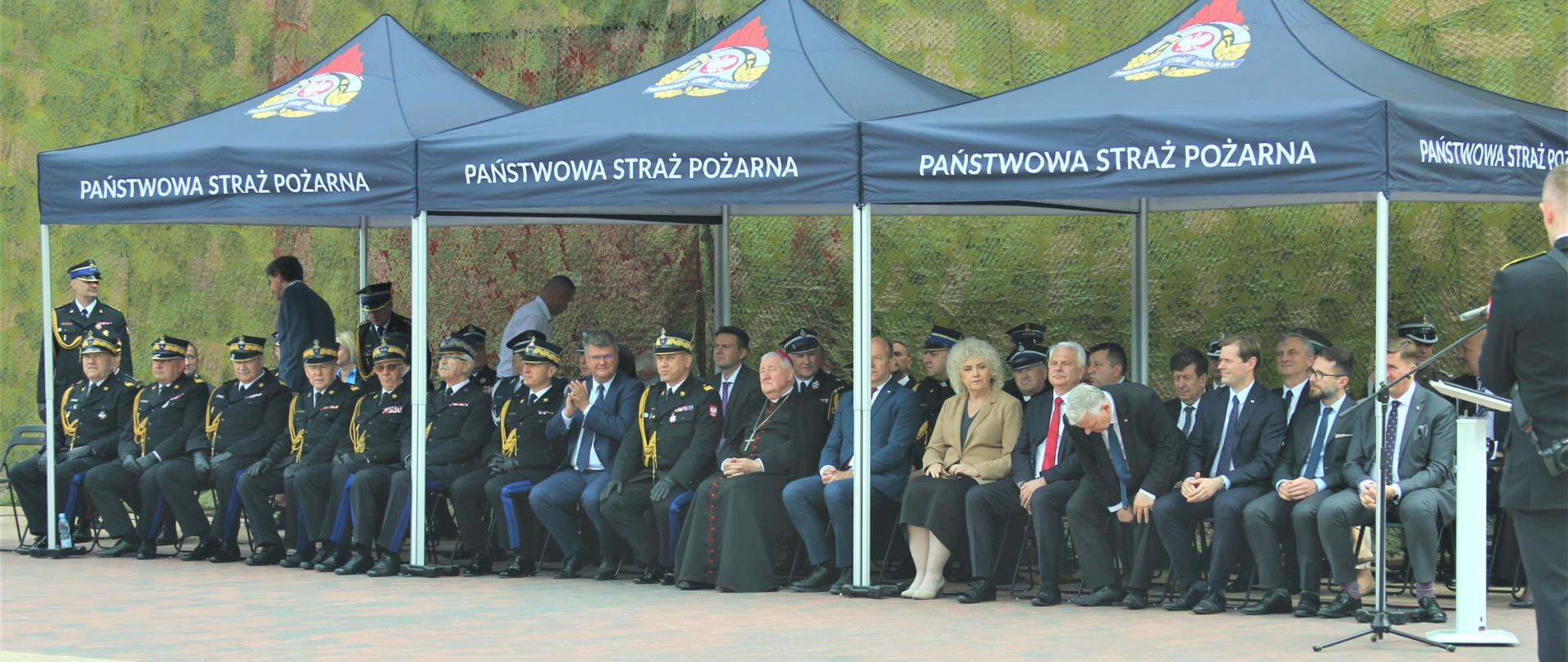 Na zdjęciu trybuna honorowa a na niej zaproszeni goście siedzący na krzesłach pod namiotami z logo i napisem Państwowa Straż Pożarna.