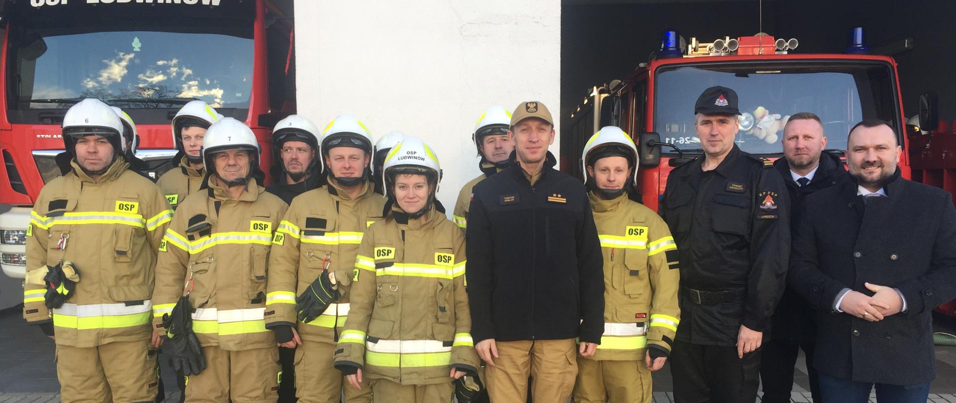 grupa strażaków w jasnych mundurach i w białych hełmach oraz strażak w czarnym mundurze w czapeczce i dwóch mężczyzn w garniturach stoją przed dwoma samochodami pozarniczymi które stoją w boksach garażowych