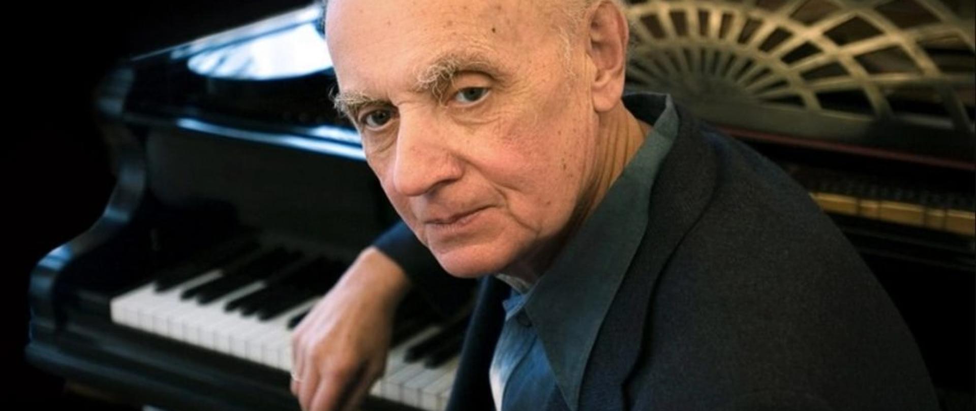 zdjęcie przedstawia starszego mężczyznę siedzącego tyłem do klawiatury fortepianu, prawe ramię wsparte na instrumencie, mężczyzna zamyślony spogląda w stronę obiektywu