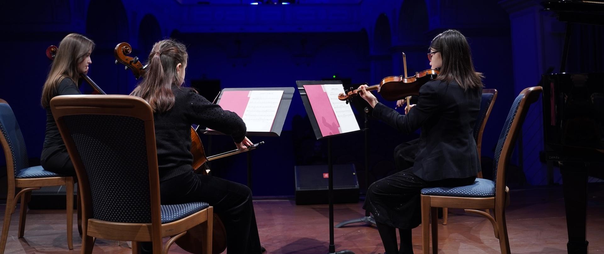 zdjęcie przedstawia trzy uczennice z kwartetu smyczkowego podczas wykonania utworu na scenie sali koncertowej, ujęcie od tyłu z widokiem na przestrzeń sali koncertowej