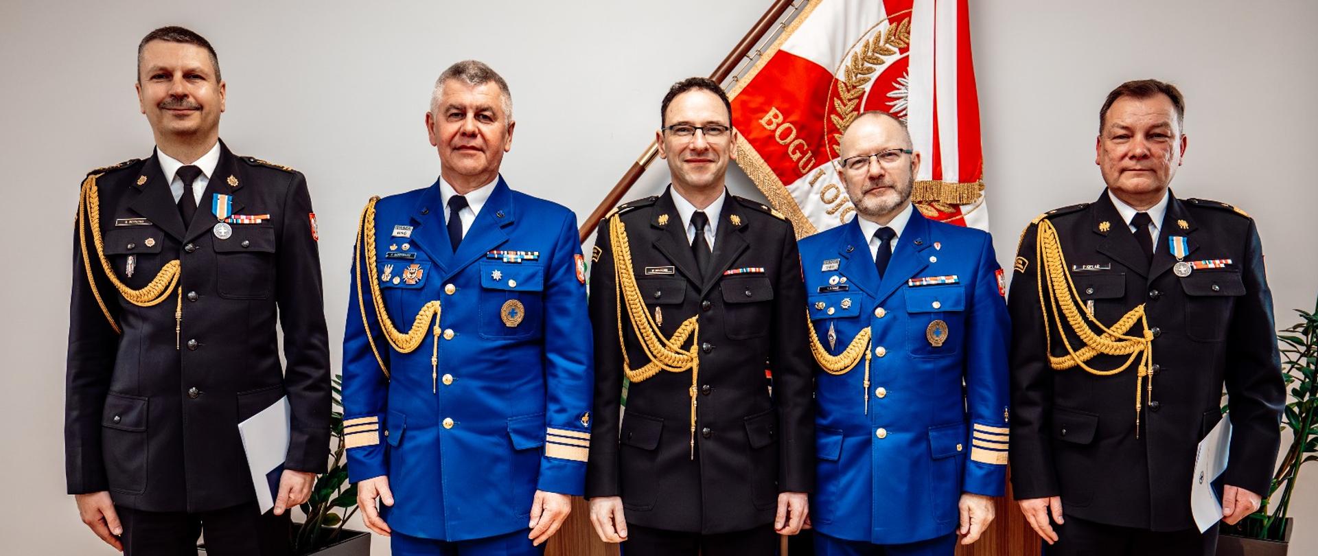 Pięciu mężczyzn, trzech w mundurach PSP i dwóch w mundurach WOPR, pozuje wspólnie do zdjęcia pamiątkowego na tle sztandaru i godła RP. 
