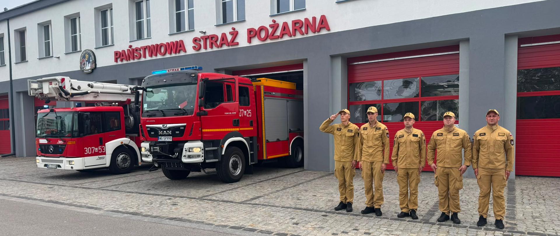 Strażacy w piaskowych mundurach oddają hołd przed garażem jednostki, obok stoją czerwone samochody z włączonymi sygnałami dźwiękowymi i świetlnymi.