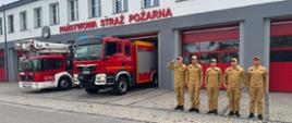 Strażacy w piaskowych mundurach oddają hołd przed garażem jednostki, obok stoją czerwone samochody z włączonymi sygnałami dźwiękowymi i świetlnymi.