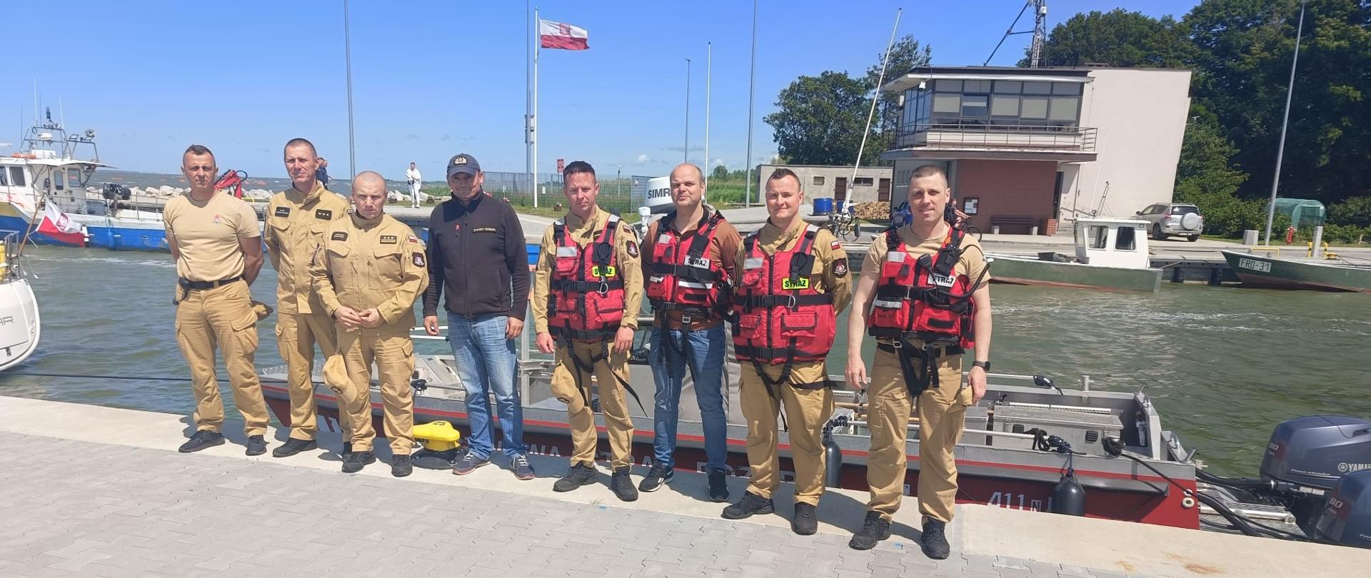 Zdjęcie przedstawia grupę mężczyzn stojących na nabrzeżu przy łodzi motorowej. Część z nich ubrana jest w mundury ratownicze lub strażackie w kolorze piaskowym, a czterech z nich ma na sobie również czerwone kamizelki ratunkowe. W tle widać inne łodzie, budynek portowy oraz powiewającą Polską flagę. Scena ma miejsce przy zbiorniku wodnym, prawdopodobnie w porcie lub przystani. Wygląda to na szkolenie lub ćwiczenia służb ratowniczych związanych z wodą.