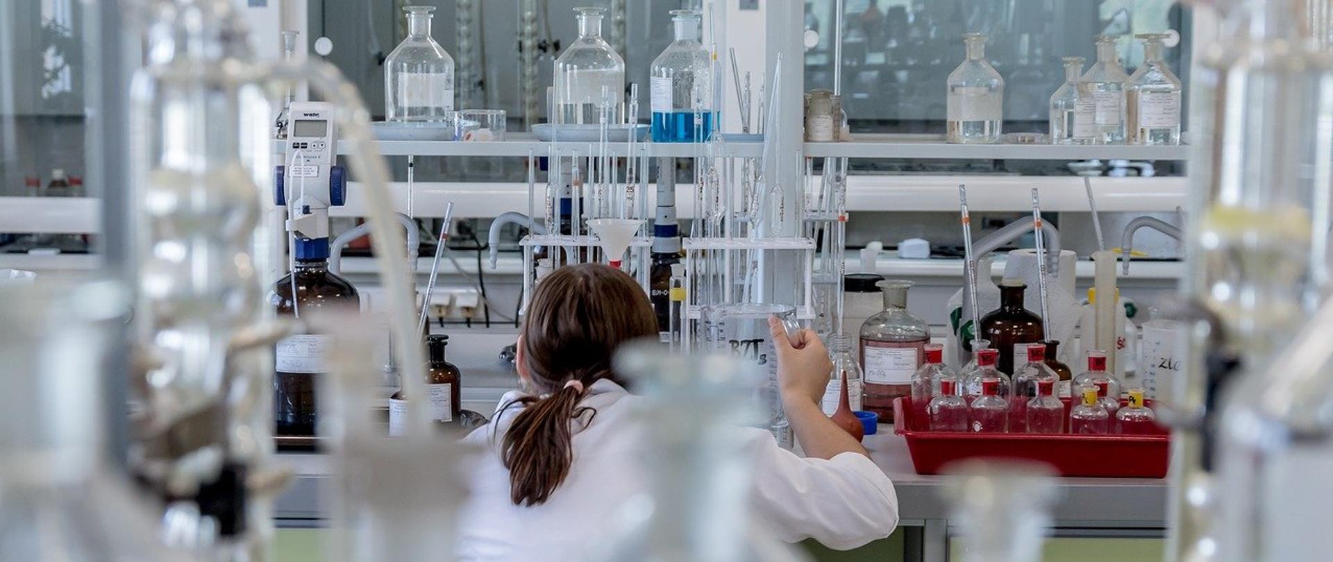 zdjęcie przedstawia laboratorium w którym przy stole znajduje się osoba wykonująca badania. na stołach widać aparaturę laboratoryjną oraz szklane butelki