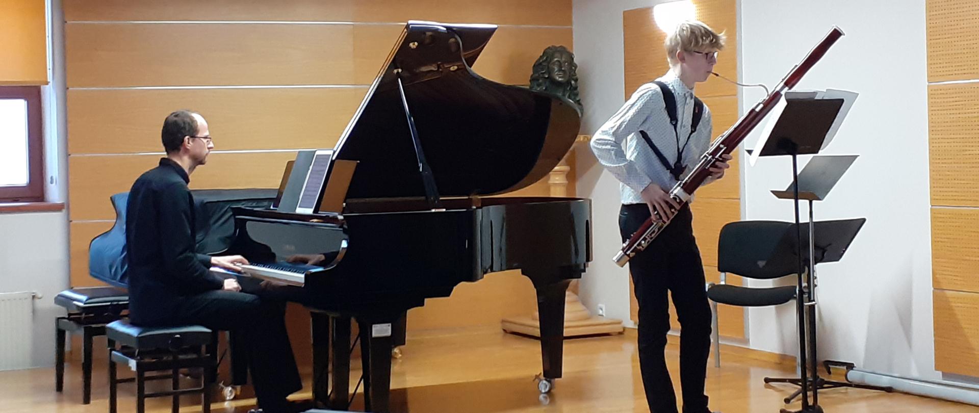 Uczeń grający na sali kameralnej, przed nim pulpit, obok krzesło. Po jego prawej stronie fortepian a przy nim akompaniator grający z nut. Na pierwszym planie z lewej stronie ksylofon. W lewym górnym rogu okno