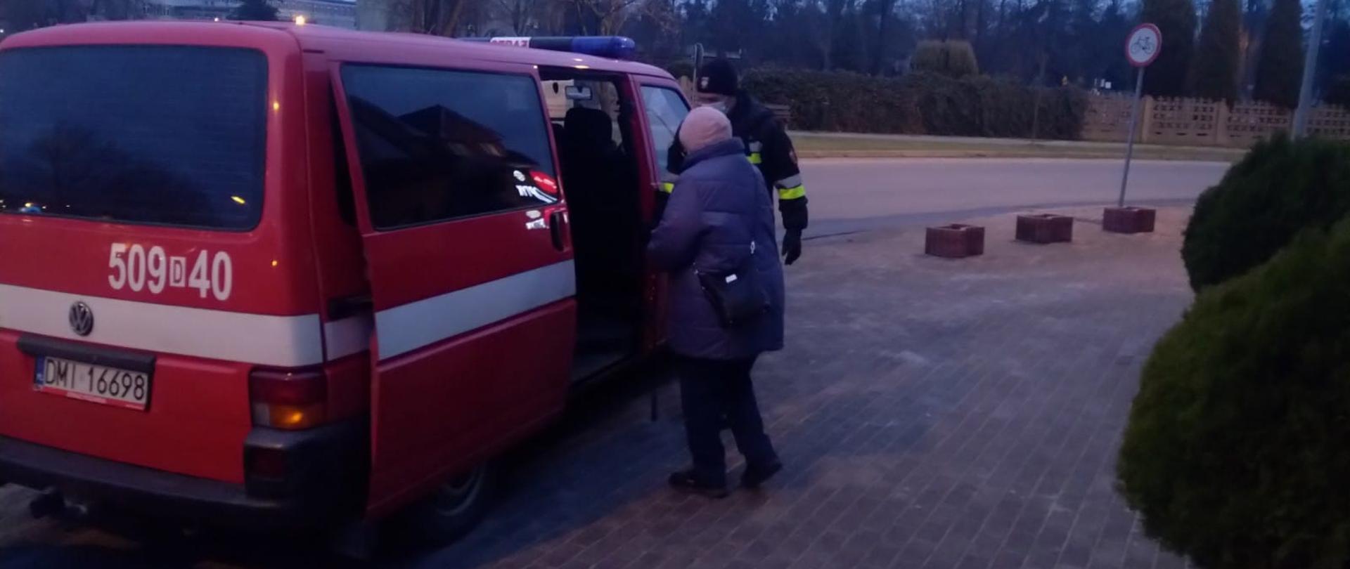 Czerwony bus straży pożarnej do którego wsiada starsza kobieta w asyście strażaka
