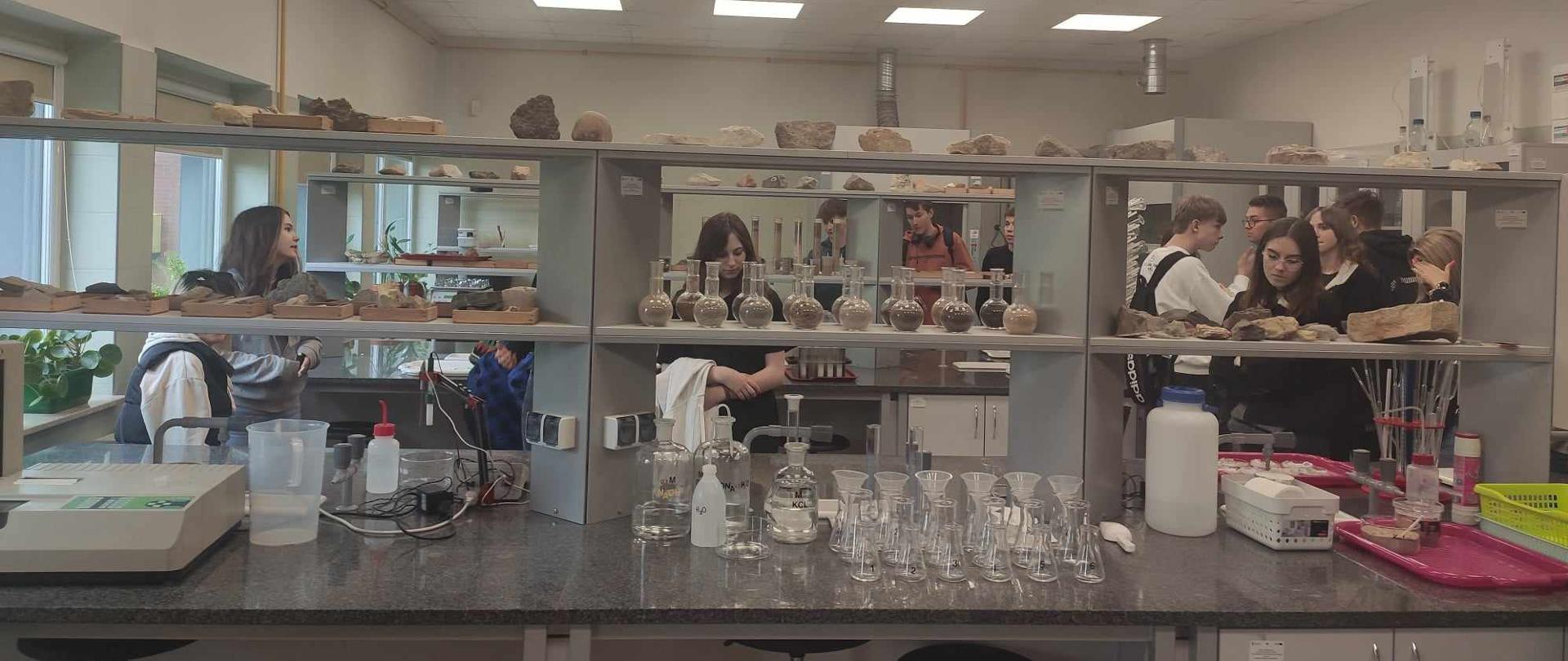 Uczniowie zwiedzają pracownię z półkami próbek skał i szkłem laboratoryjnym.