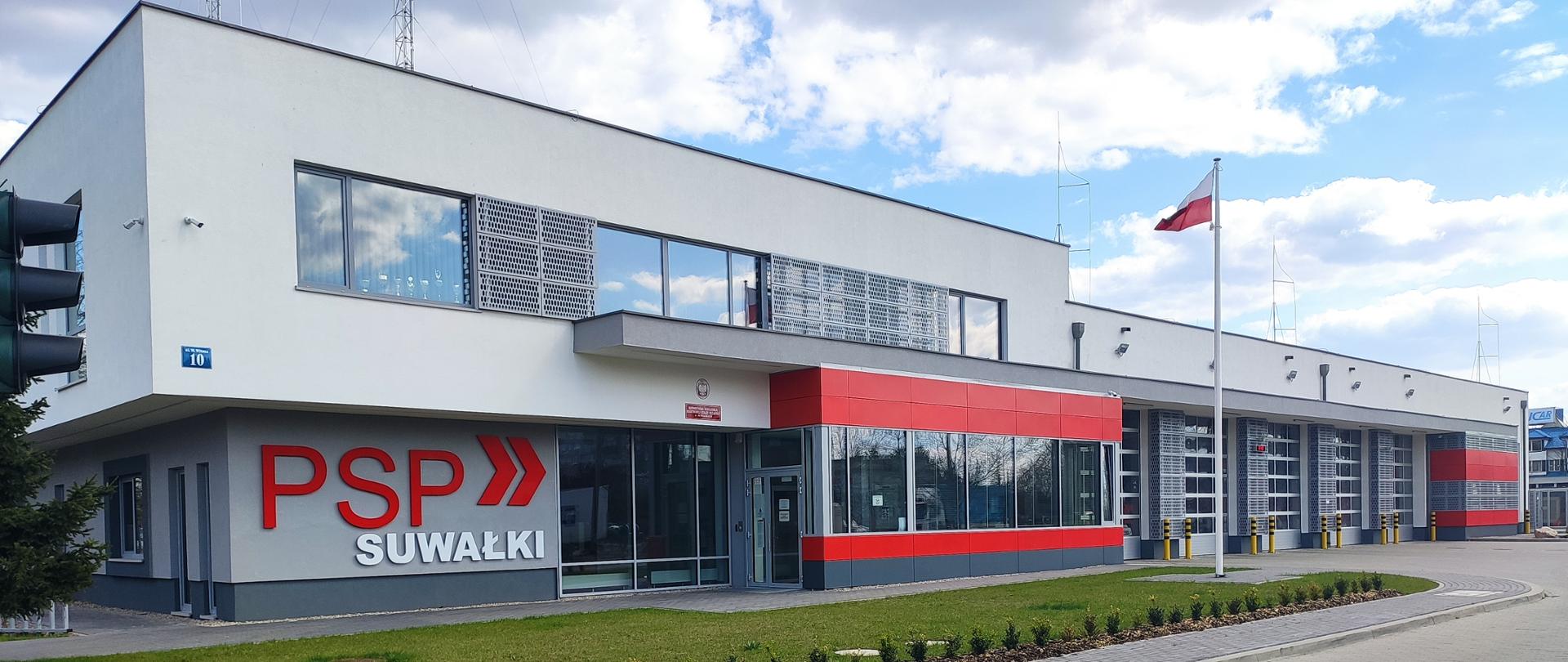 Zdjęcie obrazuje budynek KM PSP w Suwałkach na ścianie napis PSP SUWAŁKI przed budynkiem maszt z flaga państwową 