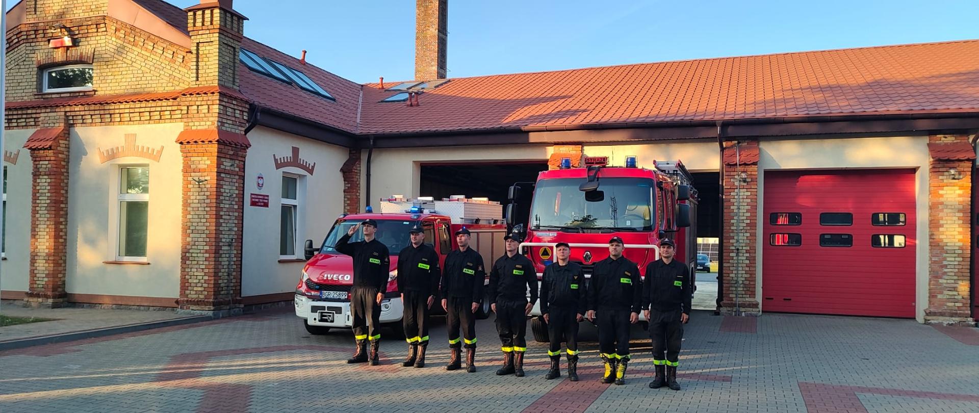 Strażacy przed budynkiem stoją w szeregu i oddają honory. W tle ssmochody strażackie.