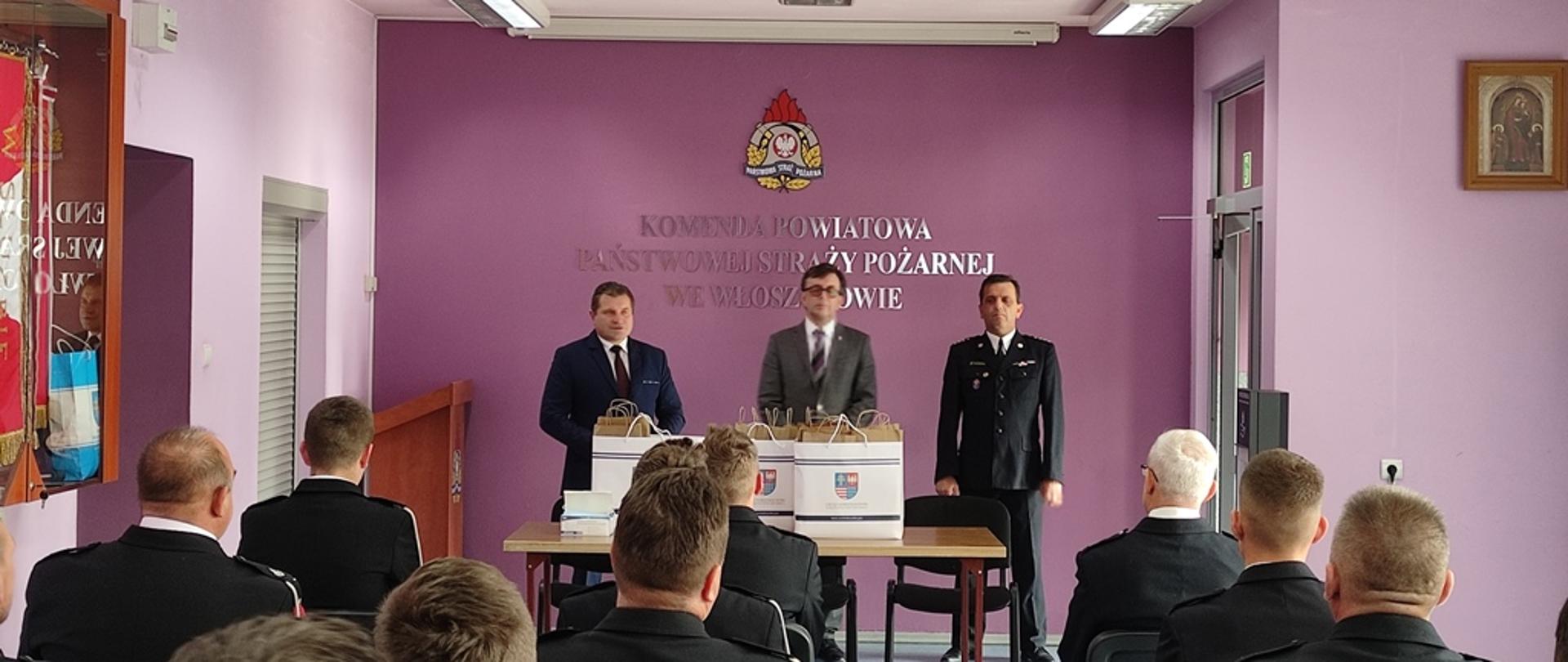 Sala Narad Komendy Powiatowej Państwowej Straży Pożarnej we Włoszczowie. Strażacy wytypowanych jednostek ochotniczych straży pożarnych z terenu powiatu włoszczowskiego ubrani w mundury galowe z sznurem otrzymują z rąk radnego sejmiku pulsoksymetry