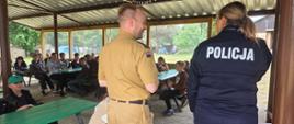 Strażak i policjantka stoją przodem do grupy dzieci i młodzieży siedzących przy stołach pod zadaszoną wiatą podczas spotkania edukacyjnego na terenie leśnego obozu.