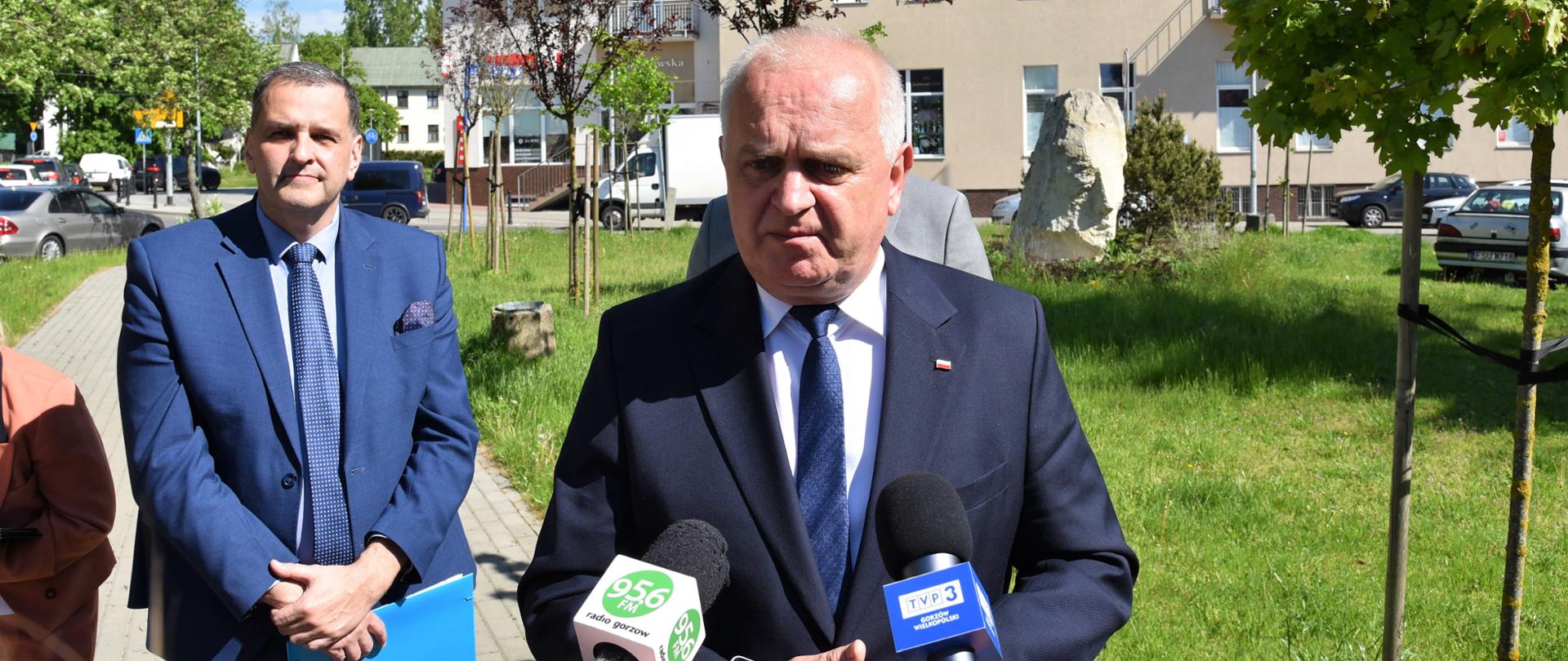 Na pierwszym planie wojewoda mówi do dziennikarzy trzymających wyciągnięte mikrofony. Za nim w tle pozostali uczestnicy konferencji, trawiasty kawałek skweru i budynek. 