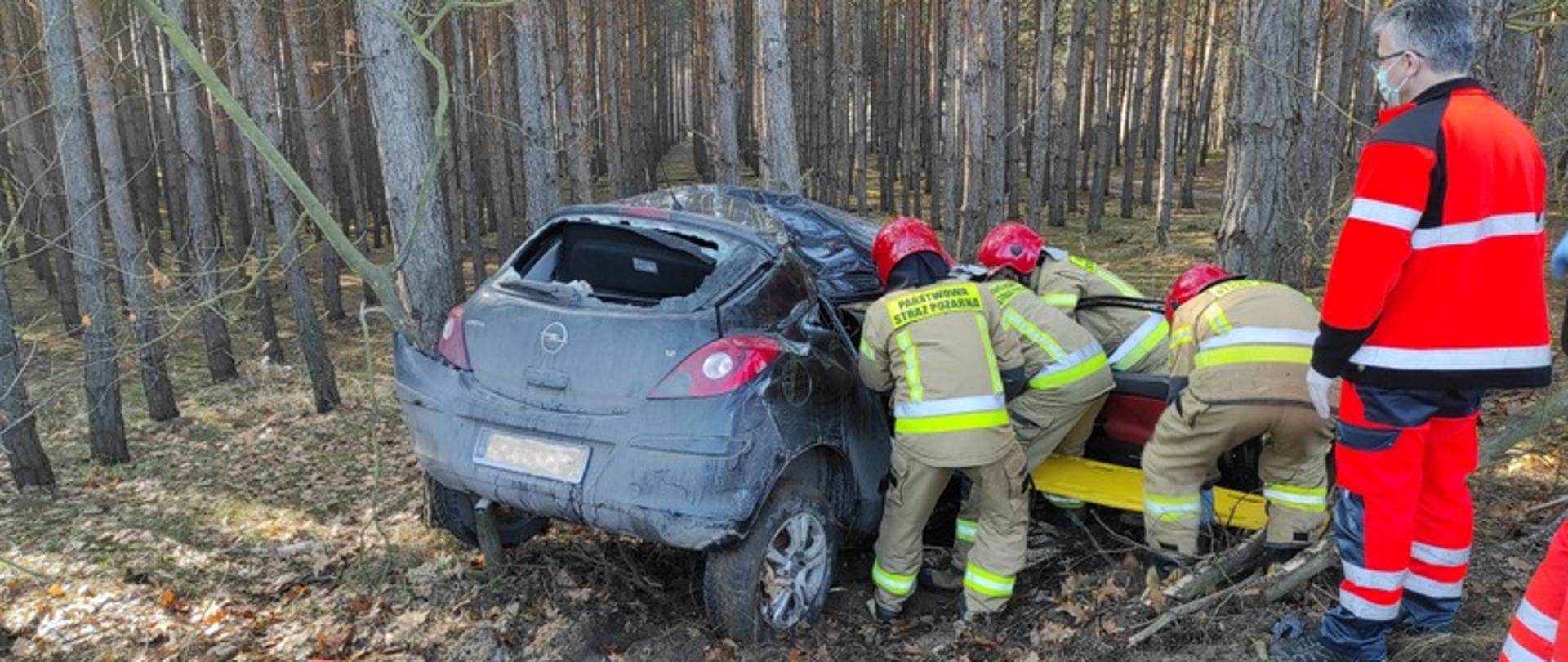 Na zdjęciu widać ciemny samochód Opel Corsa, który wjechał do lasu, uderzając w drzewo. Od strony pasażera, czterech strażaków wydobywa poszkodowaną pasażerkę auta.