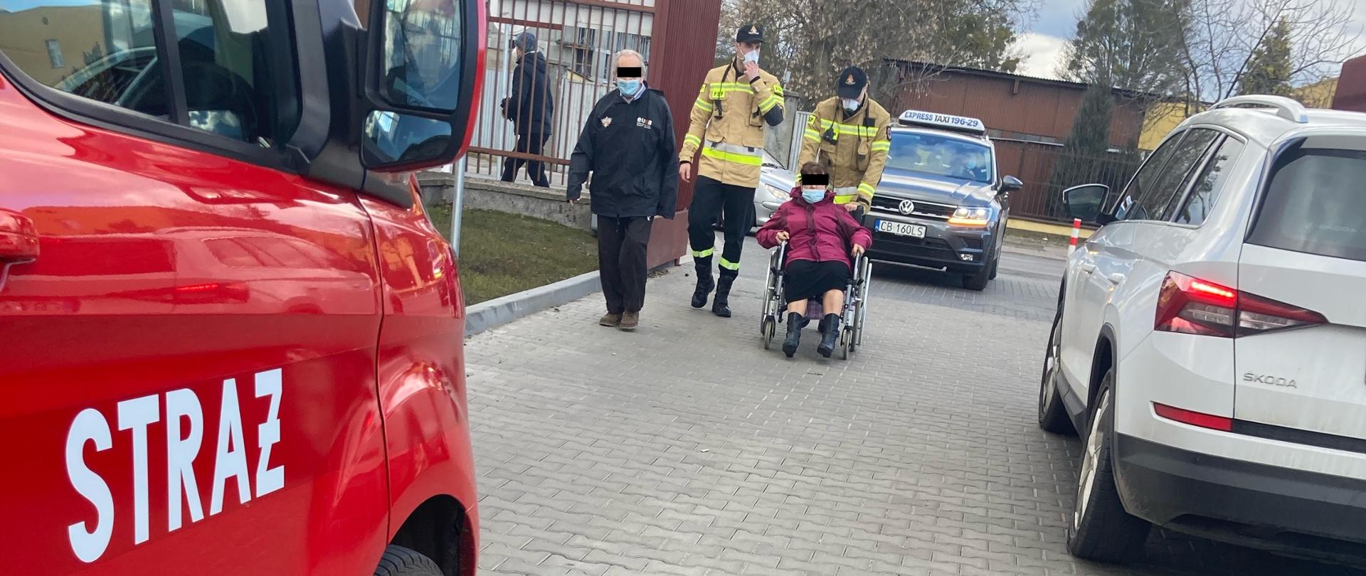 Z lewej na pierwszym planie widać prawe przednie drzwi busa strażackiego koloru czerwonego z białym napisem Straż, w tle dwóch strażaków ubranych w kurtkę ubrania specjalnego i czarne spodnie koszarowe. Jeden ze strażaków pcha wózek inwalidzki, na którym siedzi kobieta. Obok strażaków idzie mężczyzna w czarnej kurtce.