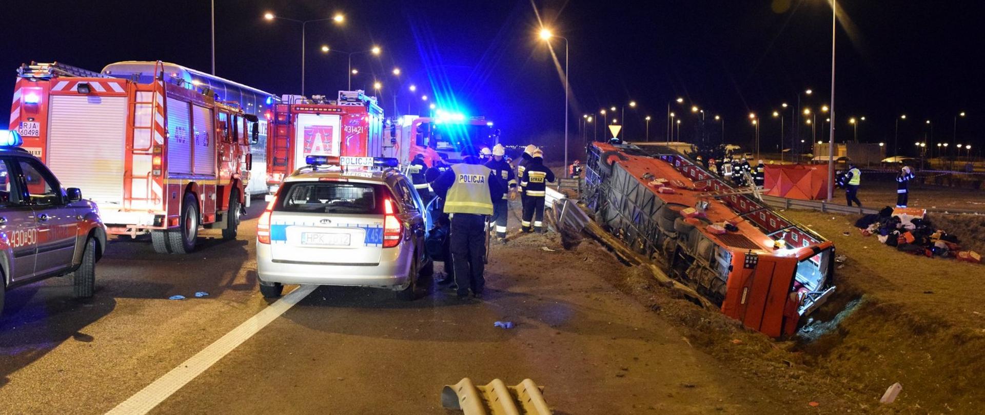 Na zdjęciu widać obraz wypadku po prawej stronie w rowie leży rozbity autokar na środku zdjęcia widać strażaków prowadzących działania ratownicze po lewej stronie widoczne samochody strażackie oraz radiowóz policyjny.