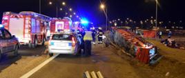 Na zdjęciu widać obraz wypadku po prawej stronie w rowie leży rozbity autokar na środku zdjęcia widać strażaków prowadzących działania ratownicze po lewej stronie widoczne samochody strażackie oraz radiowóz policyjny 