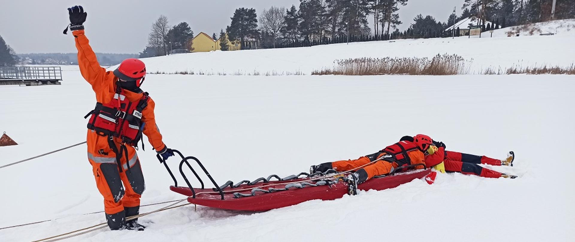 Na zdjęciu widać trzech ratowników na zamarzniętym akwenie wodnym. Zamarznięty akwen pokryty jest warstwą śniegu. Na pierwszym planie zdjęcia widać jak jeden z ratowników trzyma jedną ręką uchwyt sani lodowych natomiast drugą ma uniesioną w górę. Drugi z ratowników leży na saniach lodowych i podejmuje tonącego z przerębla. Wszyscy ratownicy ubrani są w pomarańczowe kombinezony i kaski. W tle widać linię brzegową porośniętą trzciną. Widoczne są również w oddali pojedyncze budynki mieszkalne oraz drzewa.