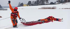 Na zdjęciu widać trzech ratowników na zamarzniętym akwenie wodnym. Zamarznięty akwen pokryty jest warstwą śniegu. Na pierwszym planie zdjęcia widać jak jeden z ratowników trzyma jedną ręką uchwyt sani lodowych natomiast drugą ma uniesioną w górę. Drugi z ratowników leży na saniach lodowych i podejmuje tonącego z przerębla. Wszyscy ratownicy ubrani są w pomarańczowe kombinezony i kaski. W tle widać linię brzegową porośniętą trzciną. Widoczne są również w oddali pojedyncze budynki mieszkalne oraz drzewa.