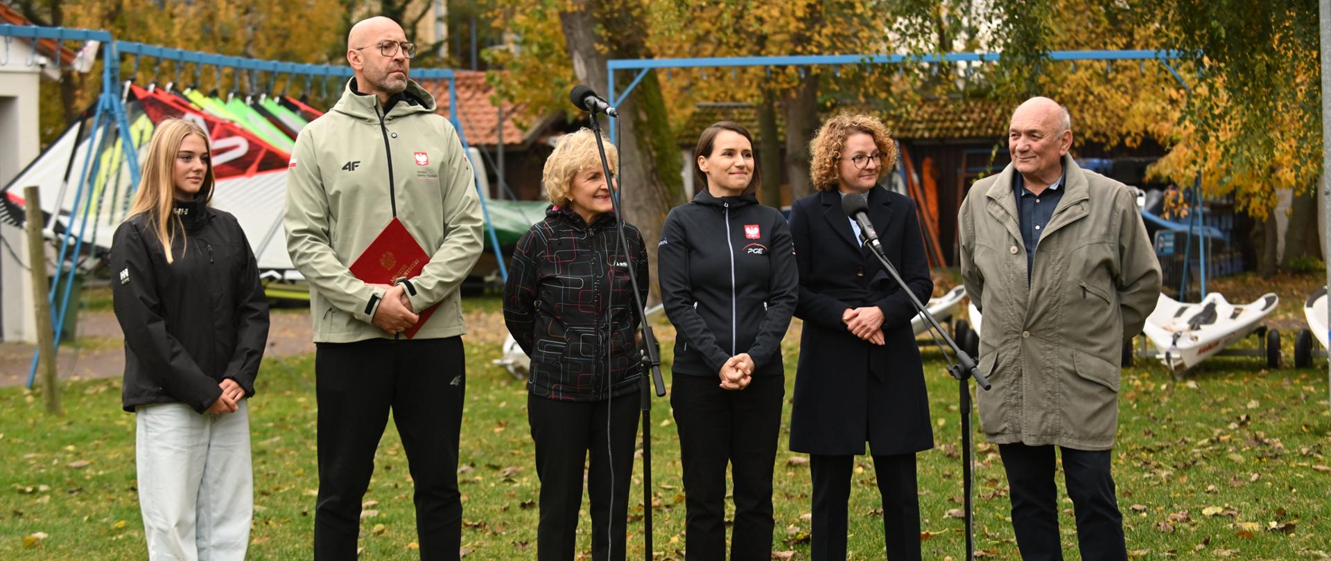 Zdjęcie. Uczestnicy konferencji stoją na trawniku. Od lewej stoją: Maria Miarczyńska, mistrzyni Europy do lat 15 w klasie iQFOiL, minister sportu i turystyki Jakub Rutnicki, posłanka Magdalena Kołodziejczak, posłanka Agnieszka Pomaska, prezydent Sopotu Magdalena Czarzyńska-Jachim i komandor Piotr Hlavaty.Przed nimi mikrofony. W tle widać ułożone jeden obok drugiego żagle, drzewa i zabudowę.