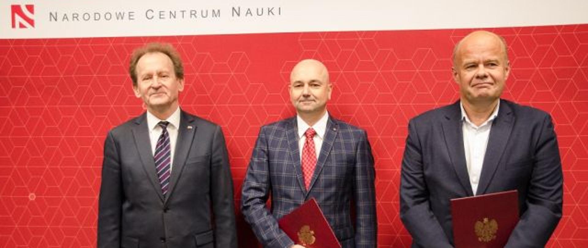 Na tle czerwonej ściany z napisem Narodowe Centrum Nauki stoi wiceminister Bernacki i dwóch mężczyzn w garniturach.