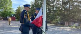 Strażacy w wyjściowych mundurach wieszają flagę państwową.