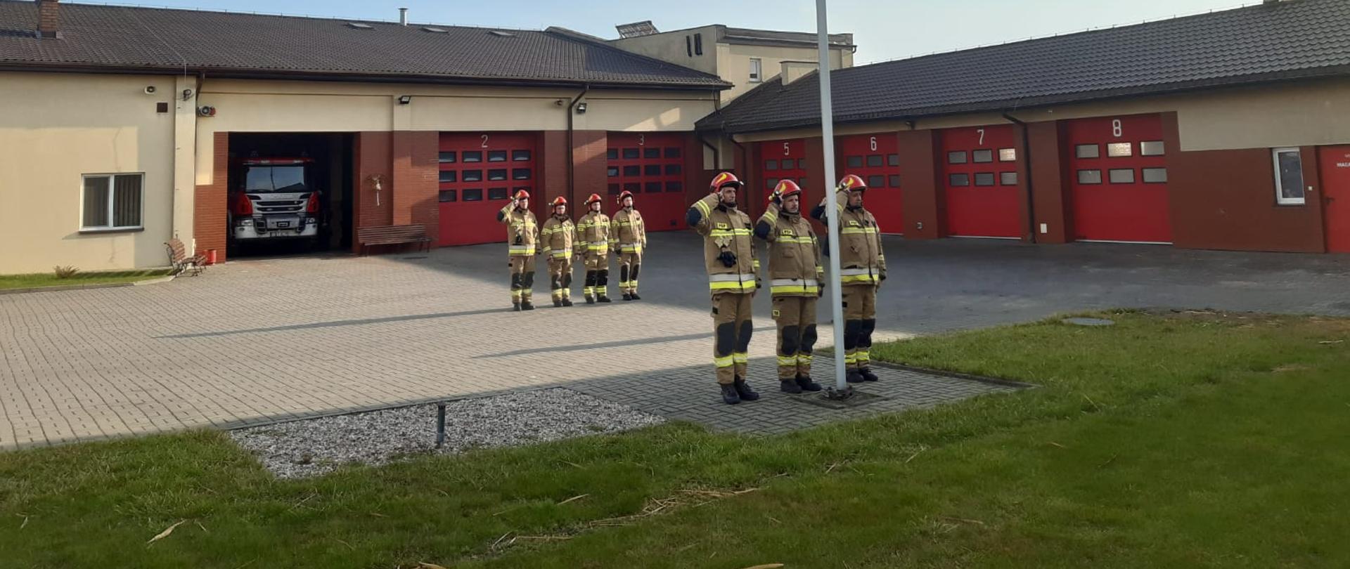Na zdjęciu widzimy ośmiu strażaków znajdujących się na placu Komendy Powiatowej Państwowej Straży Pożarnej w Rypinie, którzy oddają honor w stronę flagi państwowej. W tle widać garaże i wóz strażacki.