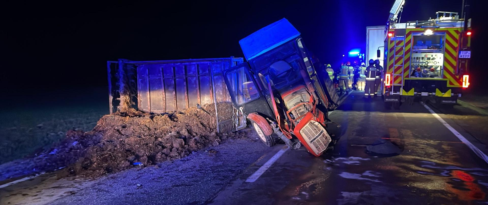Przewrócony ciągnik z rozrzutnikiem do obornika. Na drodze wysypany obornik. W tle samochody pożarnicze