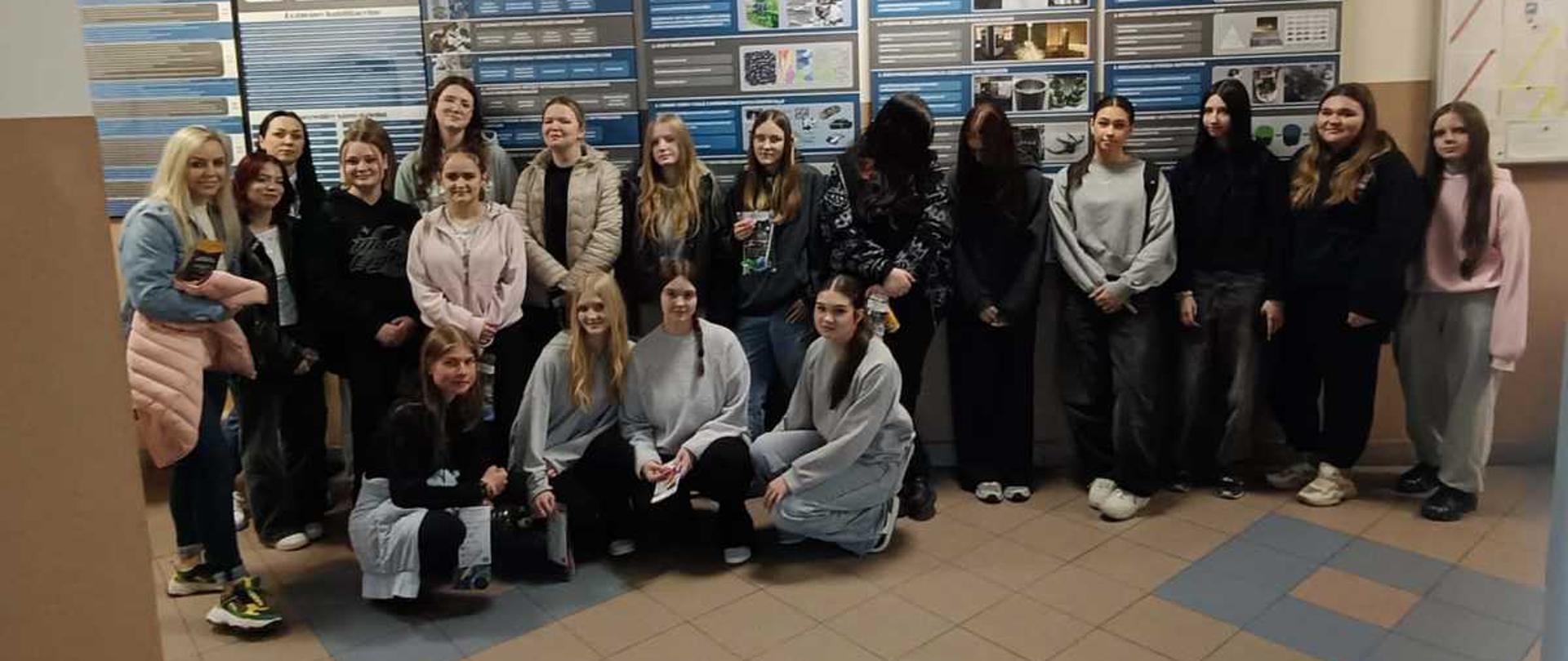 Na zdjęciu widoczna jest grupa młodych osób, ustawionych do wspólnego zdjęcia wewnątrz budynku uczelni. Część osób stoi, a kilka siedzi lub kuca z przodu. Wszyscy są ubrani w codzienne, wygodne ubrania – bluzy, kurtki i spodnie sportowe. Na ścianie za nimi umieszczone są duże tablice informacyjne z tekstami, schematami i zdjęciami dotyczącymi inżynierii materiałowej. Nad tablicami znajduje się duży napis: „Instytut Inżynierii Materiałowej – Wydział Nowych Technologii i Chemii”.
