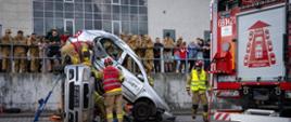 Strażacy PSP prowadzą działania ratownictwa technicznego przy dwóch uszkodzonych samochodach, obok których stoi pojazd pożarniczy, a przebieg konkurencji obserwują zgromadzeni widzowie.
