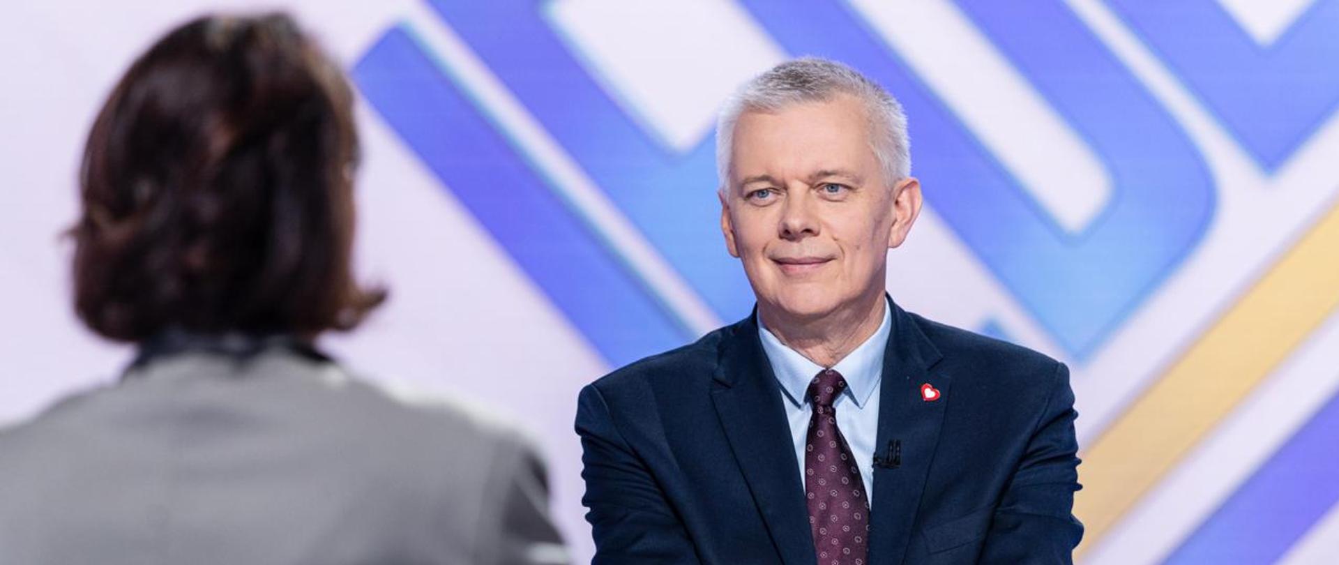 Na zdjęciu wnętrze studnia telewizyjnego. Po prawej stronie siedzi minister Tomasz Siemoniak, po lewej stronie redaktor Agata Adamek (siedzi tyłem do obiektywu). W tle widać fragment napisu z nazwą programu "JEDEN NA JEDEN" (niebieskie litery i żółta linia podkreślająca.