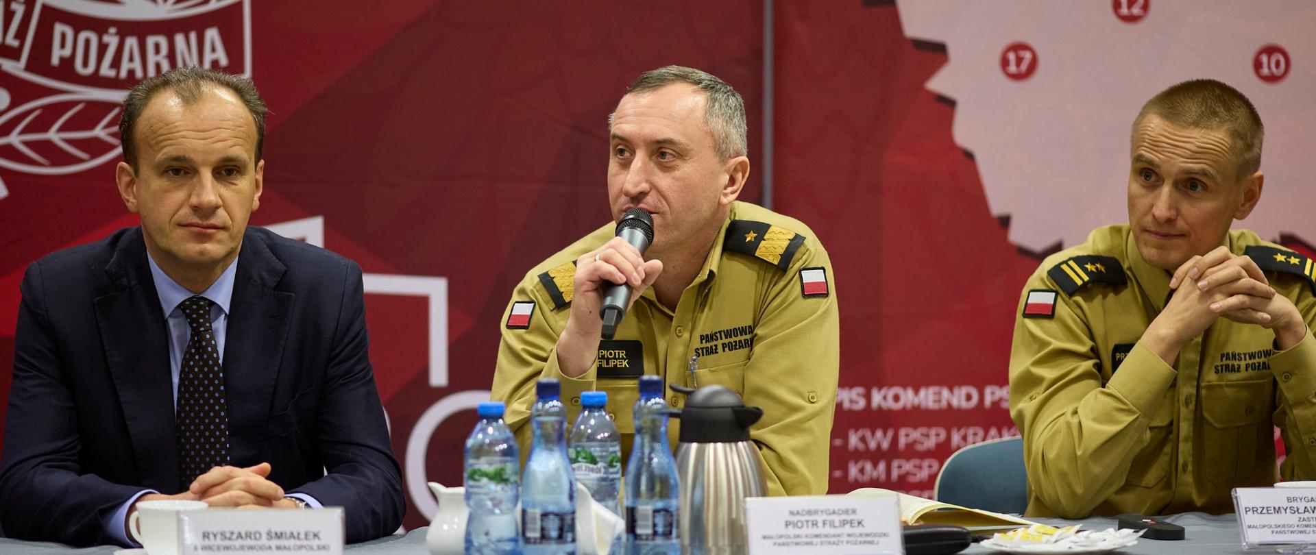 Na zdjęciu widać trzy osoby. W środku strażak z mikrofonem przy stoliku prezydialnym.