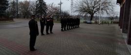 Strażacy ustawieni w rzędzie, poczet flagowy oraz dowódca przed budynkiem Komendy.