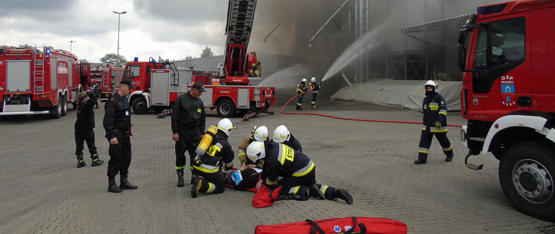 na zdjęciu strażacy udzielający pomocy poszkodowanemu w tle samochody ciężarowe straży oraz zadymione silosy.