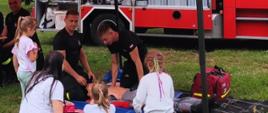 dwóch strażaków na boisku prezentuje pokaz pierwszej pomocy zgromadzonym dzieciom, w tle samochód pożarniczy