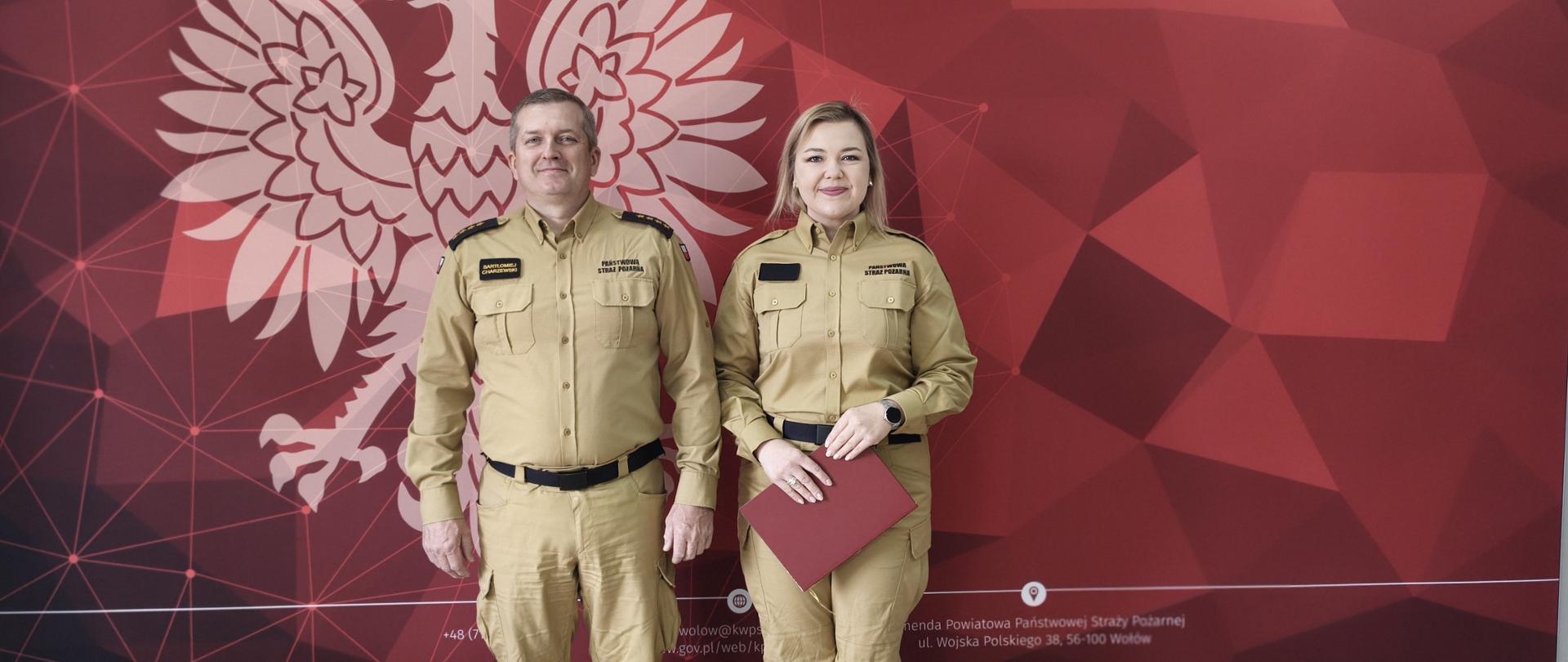 Zdjęcie pamiątkowe przedstawiające dwoje funkcjonariuszy Państwowej Straży Pożarnej na tle oficjalnej ścianki. Po lewej stronie stoi Komendant Powiatowy PSP w Wołowie, a po prawej kobieta – nowa funkcjonariuszka po złożonej rocie ślubowania. Oboje są ubrani w piaskowe mundury służbowe. Kobieta trzyma w dłoniach bordową teczkę z dokumentami. W tle znajduje się czerwona grafika z geometrycznymi wzorami, na której widnieje duże białe godło Polski (orzeł w koronie), napis „PAŃSTWOWA STRAŻ POŻARNA KOMENDA POWIATOWA PSP W WOŁOWIE” oraz logotyp straży pożarnej. Na dole ścianki umieszczono dane adresowe jednostki. Podłoga w pomieszczeniu wykonana jest z jasnych paneli imitujących drewno.