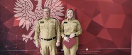Zdjęcie pamiątkowe przedstawiające dwoje funkcjonariuszy Państwowej Straży Pożarnej na tle oficjalnej ścianki. Po lewej stronie stoi Komendant Powiatowy PSP w Wołowie, a po prawej kobieta – nowa funkcjonariuszka po złożonej rocie ślubowania. Oboje są ubrani w piaskowe mundury służbowe. Kobieta trzyma w dłoniach bordową teczkę z dokumentami. W tle znajduje się czerwona grafika z geometrycznymi wzorami, na której widnieje duże białe godło Polski (orzeł w koronie), napis „PAŃSTWOWA STRAŻ POŻARNA KOMENDA POWIATOWA PSP W WOŁOWIE” oraz logotyp straży pożarnej. Na dole ścianki umieszczono dane adresowe jednostki. Podłoga w pomieszczeniu wykonana jest z jasnych paneli imitujących drewno.