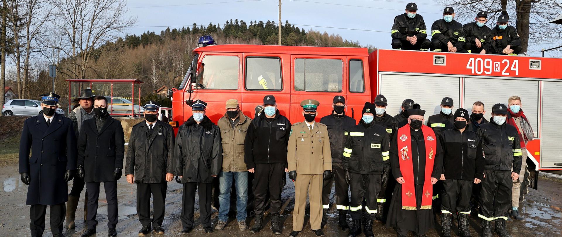 Zdjęcie przedstawia zaproszonych gości oraz strażaków na tle samochodu pożarniczego. Strażacy ubrani są w mundury galowe oraz koszarowe, wśród nich jest komendant powiatowy PSP w Ustrzykach Dolnych, ksiądz, przedstawiciel straży granicznej oraz przedstawiciele lokalnych władz. 