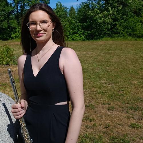 Zdjęcie kolorowe. Młoda flecistka w stroju galowym stoi z instrumentem na tle drzew.