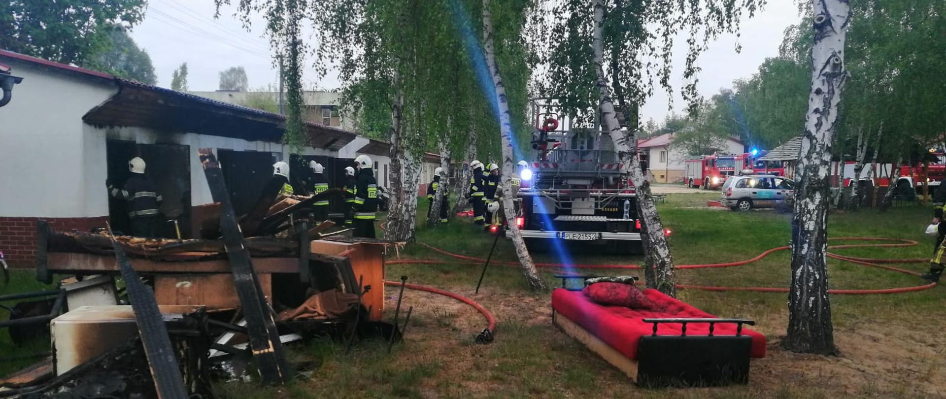 Samochód pożarniczy stoi na polanie obok strażacy przy spalonym budynku.