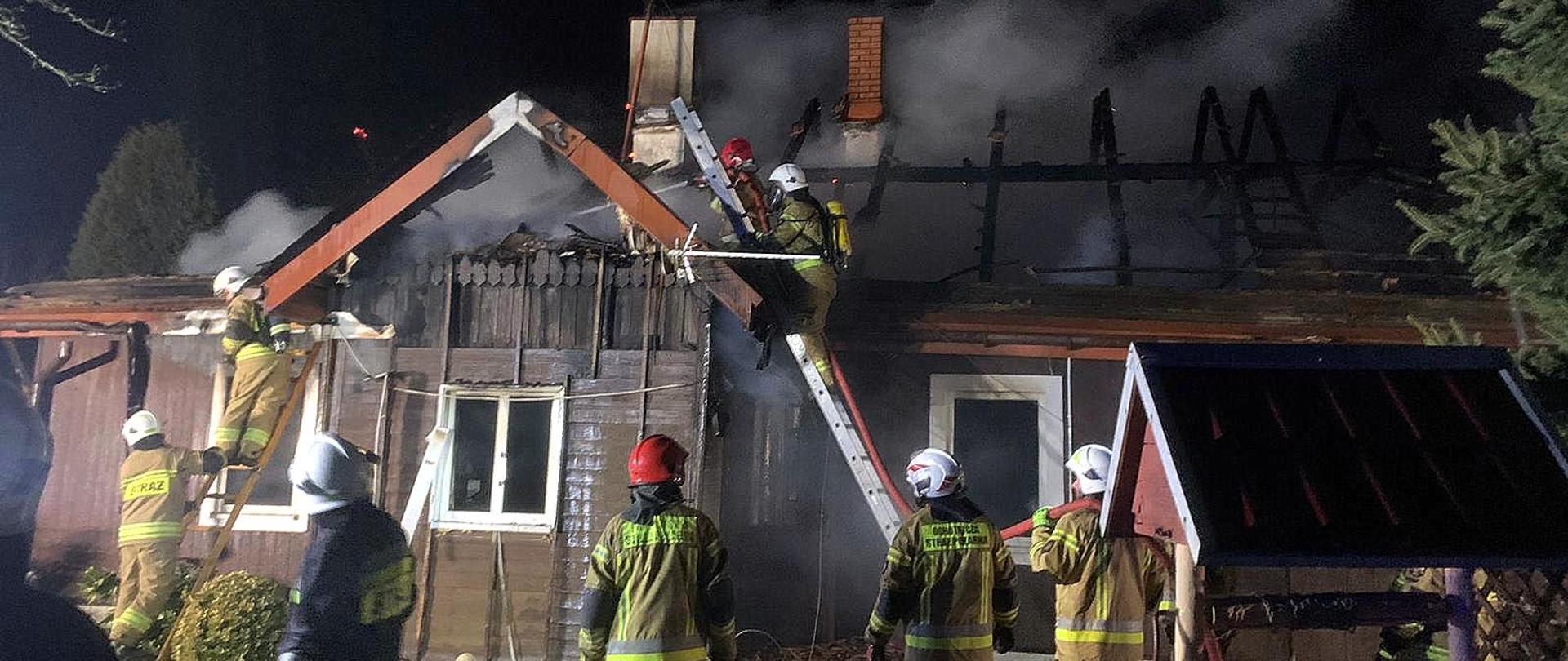 Zdjęcie zrobione na zewnątrz w nocy. Strażacy gaszą płonący dom. Czterech z nich stoi na drabinach przedstawionych do budynku gasząc pożar. Dookoła stoją strażacy uczestniczący w działaniach gaśniczych.