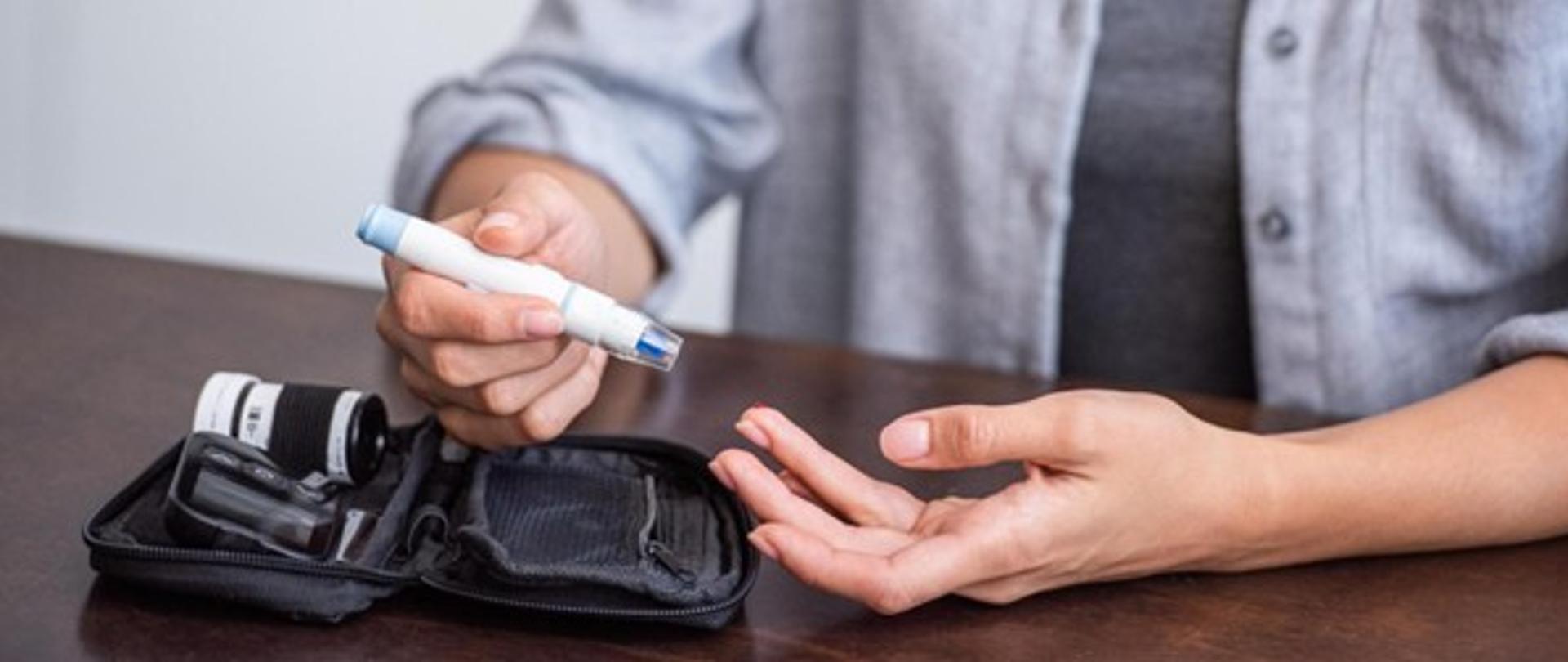 Na zdjęciu widać zbliżenie rąk osoby siedzącej przy stole. Osoba trzyma w prawej dłoni biały przyrząd z niebieską końcówką – jest to nakłuwacz do pobierania kropli krwi z palca. Lewa dłoń jest otwarta, a palec wskazujący jest przygotowany do nakłucia. Na stole leży czarne etui z zamkiem błyskawicznym, w którym znajdują się akcesoria do pomiaru poziomu cukru we krwi, w tym mały pojemnik z paskami testowymi. Tło jest neutralne, a osoba ma na sobie jasnoszarą koszulę rozpiętą na nadgarstkach. Cała scena sugeruje wykonywanie pomiaru glukozy w warunkach domowych, co jest typowe dla osób chorujących na cukrzycę.