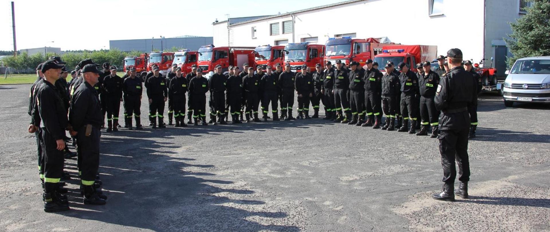 Na zdjęciu stoi grupa około 50 strażaków podczas odprawy. w tle samochody pożarnicze.