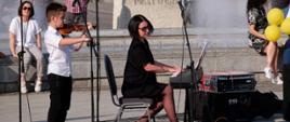 Na miejskim placu, w pobliżu fontanny i na tle pomnika widoczna kobieta grająca na fortepianie elektronicznym i chłopiec grający na skrzypcach. W tle publiczność.