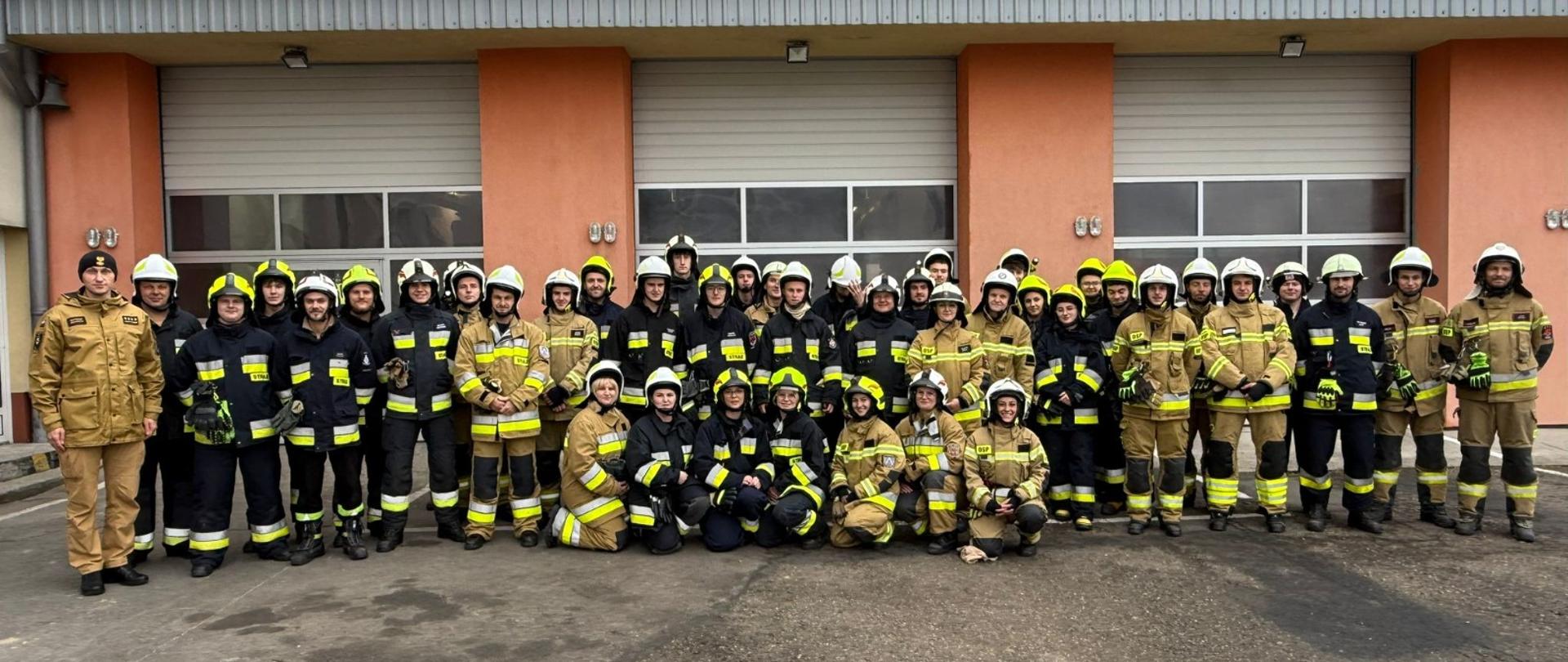 Strażacy ustawieni w kilku szeregach stoją ustawieni w na tle garażów. Ubrani są w ubrania specjalne i hełmy