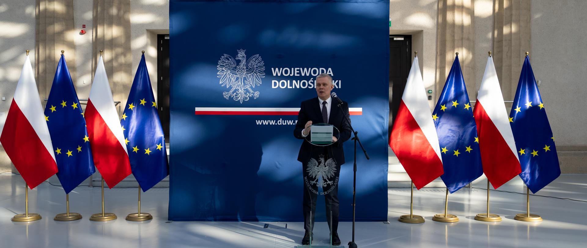 Minister Spraw Wewnętrznych i Administracji Tomasz Siemoniak na tle banneru Wojewody Dolnośląskiego. Po obu stronach banneru naprzemiennie stoją flagi Polski i Unii Europejskiej. 